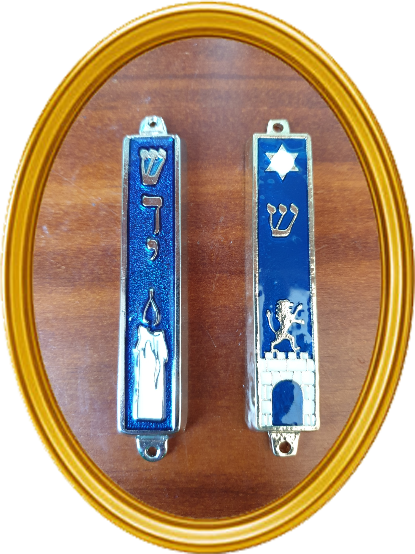 Two mezuzot on a wooden background in an oval cutout shape surrounded by a gold frame. The mezuzah on the left is silver and blue with the Hebrew word 'shaddai' and a picture of a white candle. The mezuzah on the right is gold and blue, with the Hebrew letter 'shin' and a picture of a gold lion on a white brick wall.